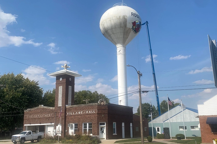 A picture of part of the city of Roseville, Illinois. The Roseville, Illinois water tower is being pressure washed by a staff member using a blue lift in the background.